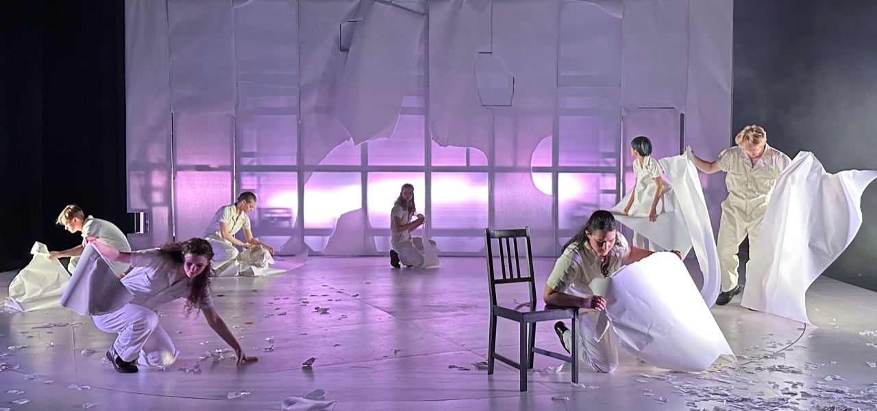 A lavender lit stage with torn white paper backdrop. Diverse young adult performers wearing white examine large scraps of paper.