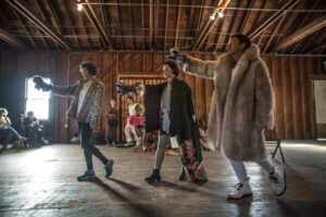 Two women and a man dressed in fur coats walk diagonally through a barn wielding raccoon puppets while a small audience looks on.