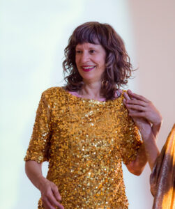 A 50 year-old white woman in a gold top and red lipstick smiles over one shoulder holding a dancer’s hand on her other shoulder.