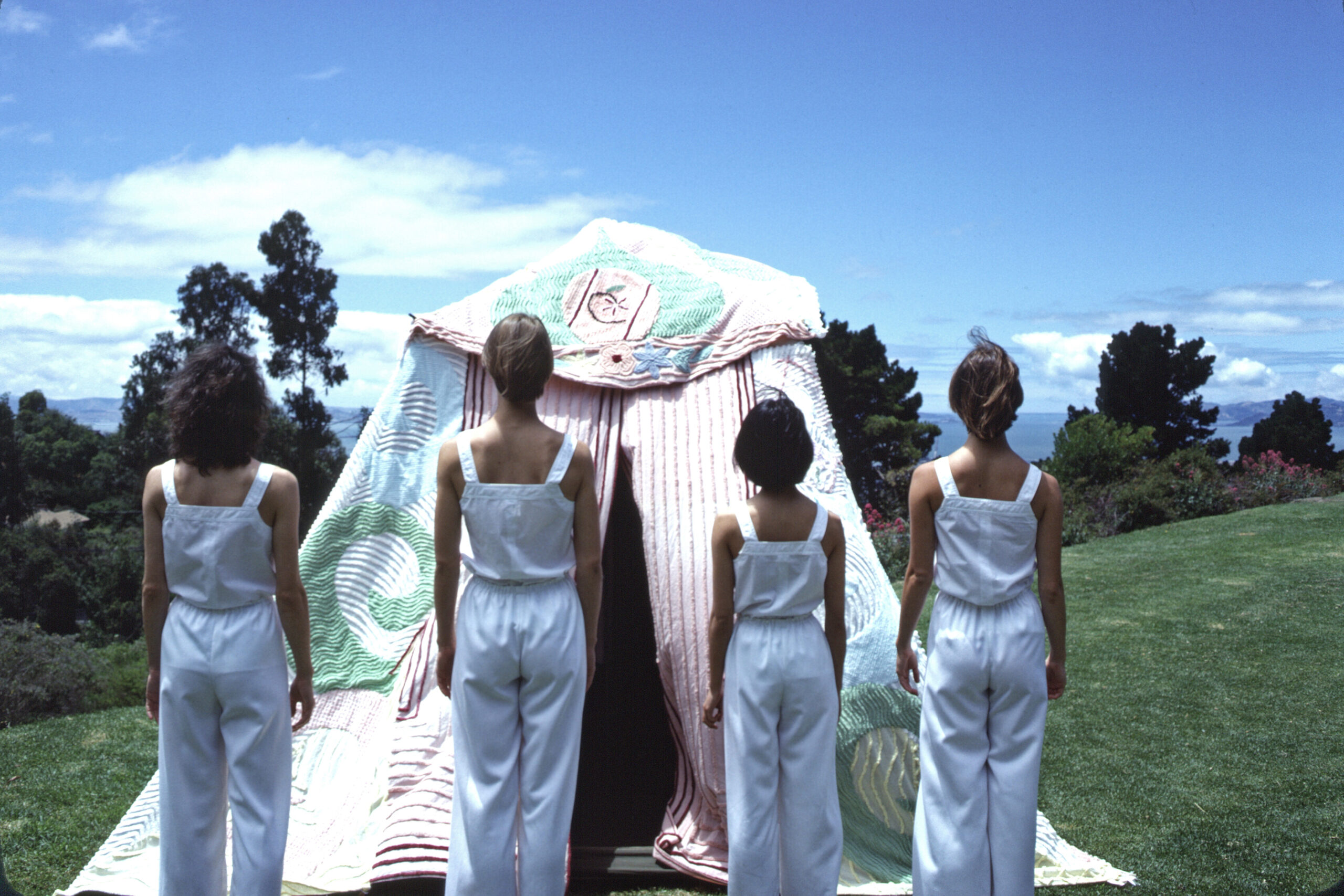 Four women, with their backs to us, look at and beyond a tall tent made out of pastel colored chenille, against a tree-lined hill.