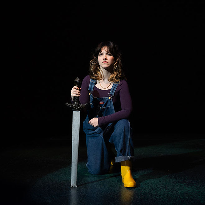 A young woman with shaggy brunette hair & blonde highlights kneels on stage with a sword. She wears a necklace, purple long sleeve, overalls, and yellow rain boots.