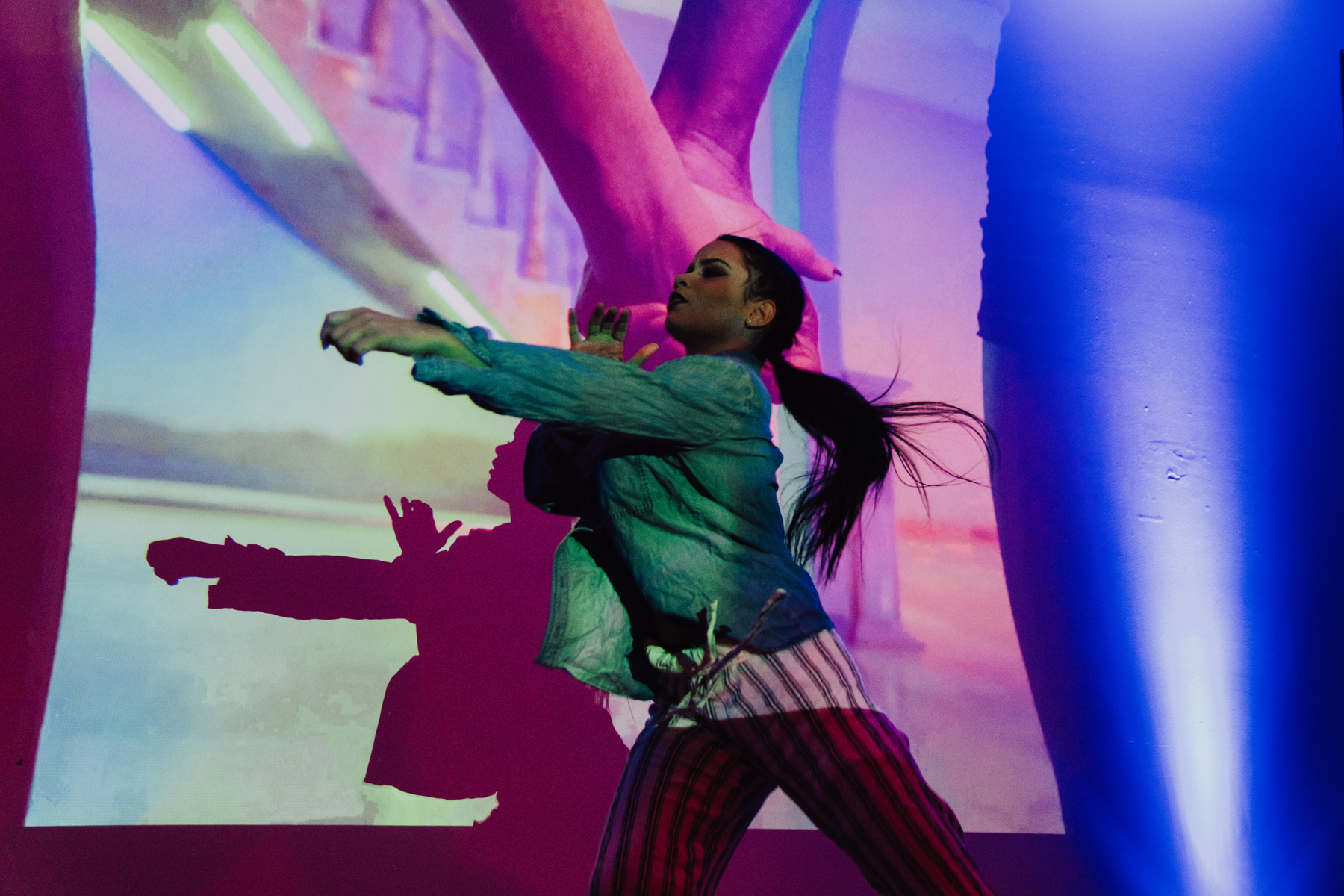 A dancer in a green long-sleeved jacket and black and white striped pants, reaches across her body as her long ponytail moves behind her.  She is in shadowy and colorful lighting in front of a video image of two holding hands.