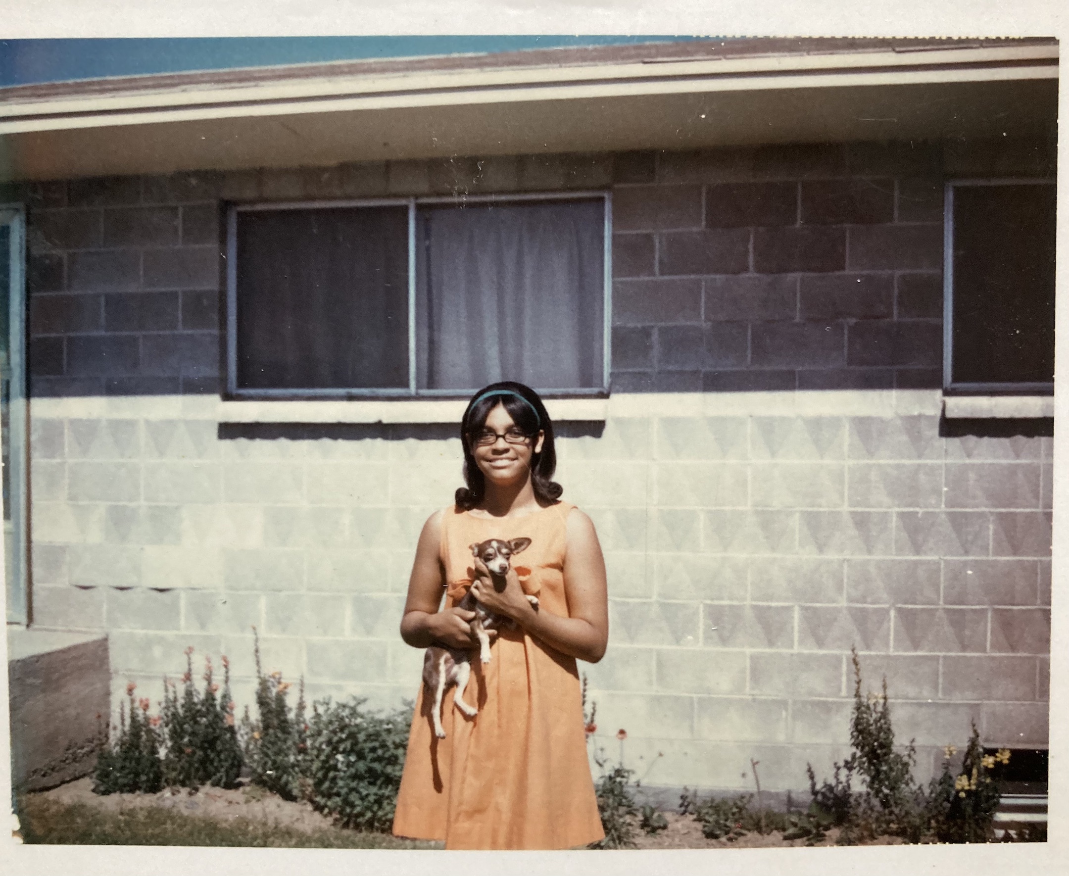 Polaroid photo of teenage black girl holding a small brown dog in front of a cinder block house in the sunlight.