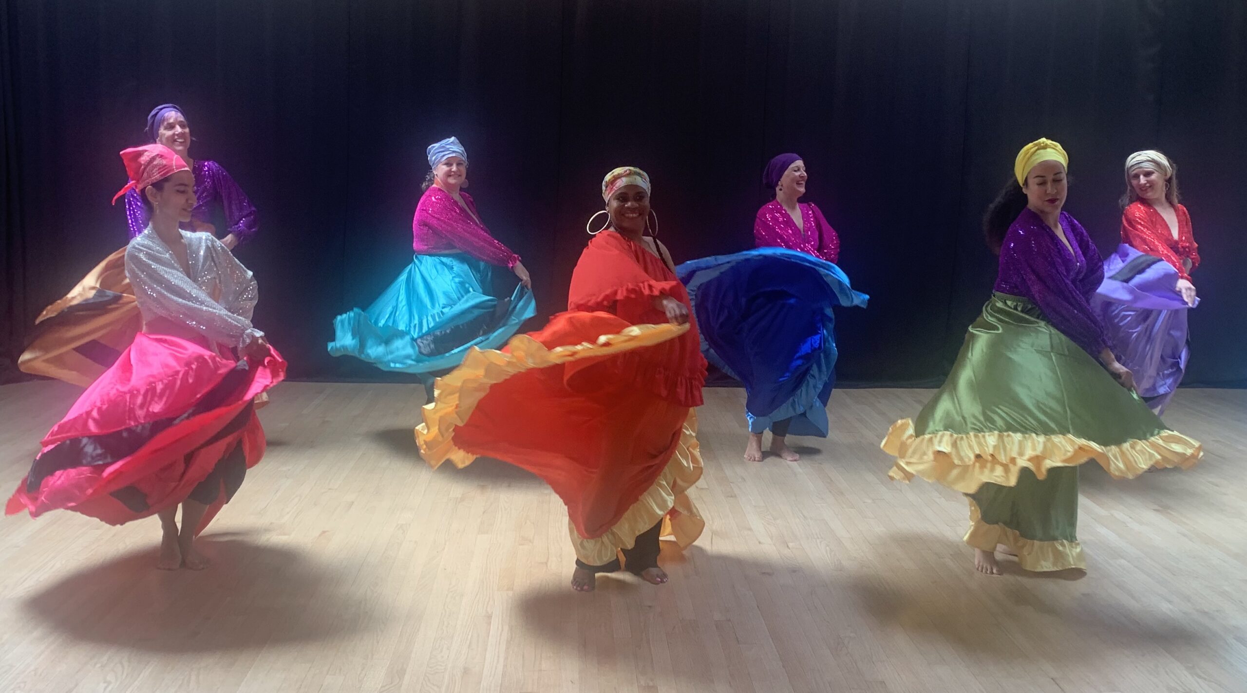 Nine dancers in brightly colored skirts and head wraps dancing smile and spin