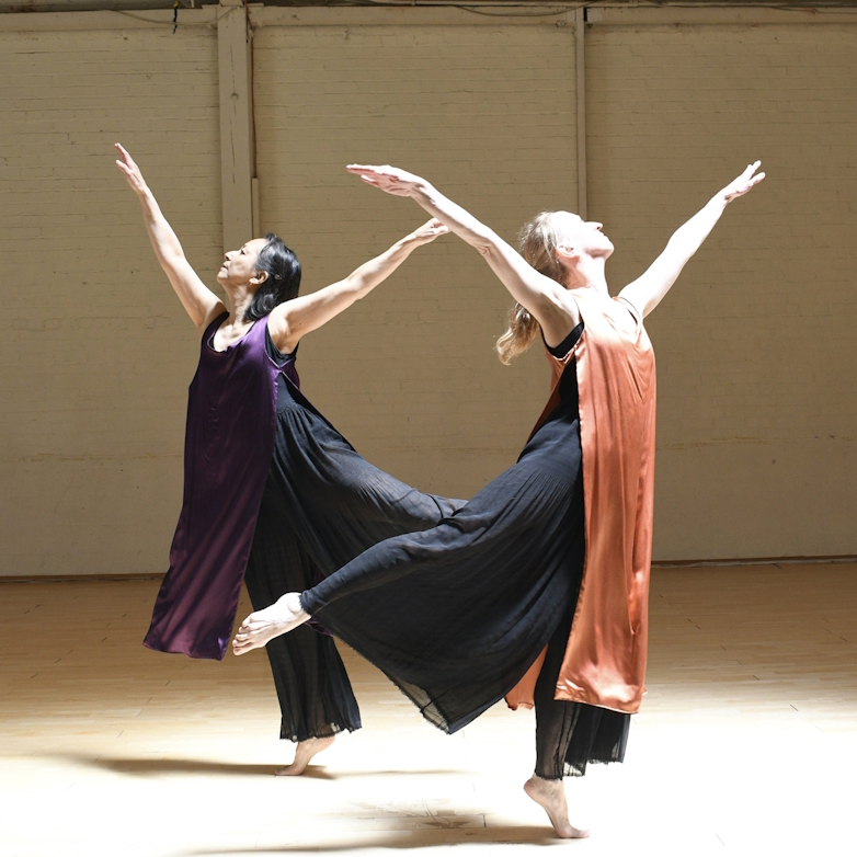 Two dancers, wearing colorful tunics move joyfully with outstretched arms on a wooden dance floor.