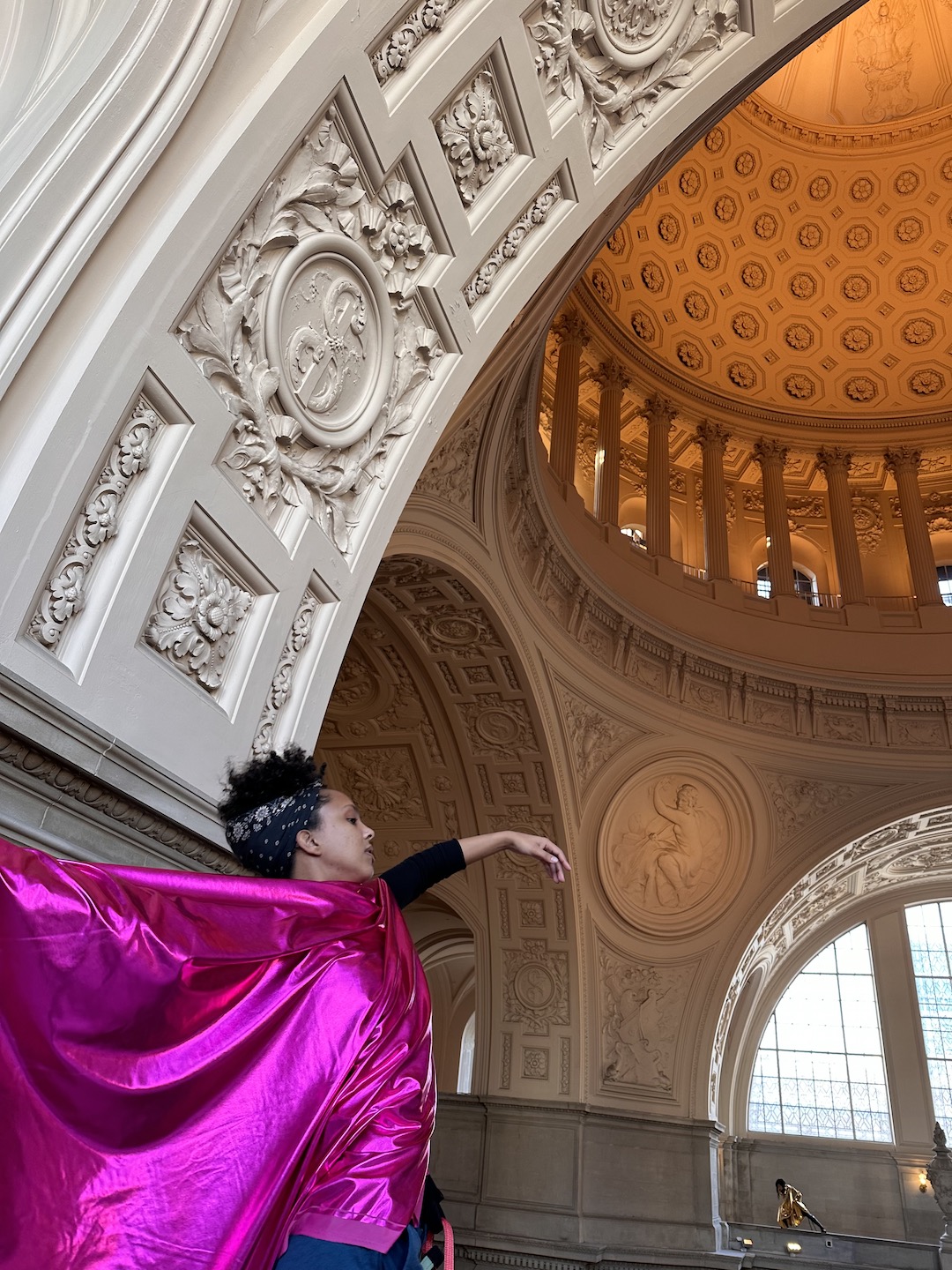 A caramel-colored Black woman draped in shiny fuchsia fabric gestures gracefully amidst neoclassical architecture.