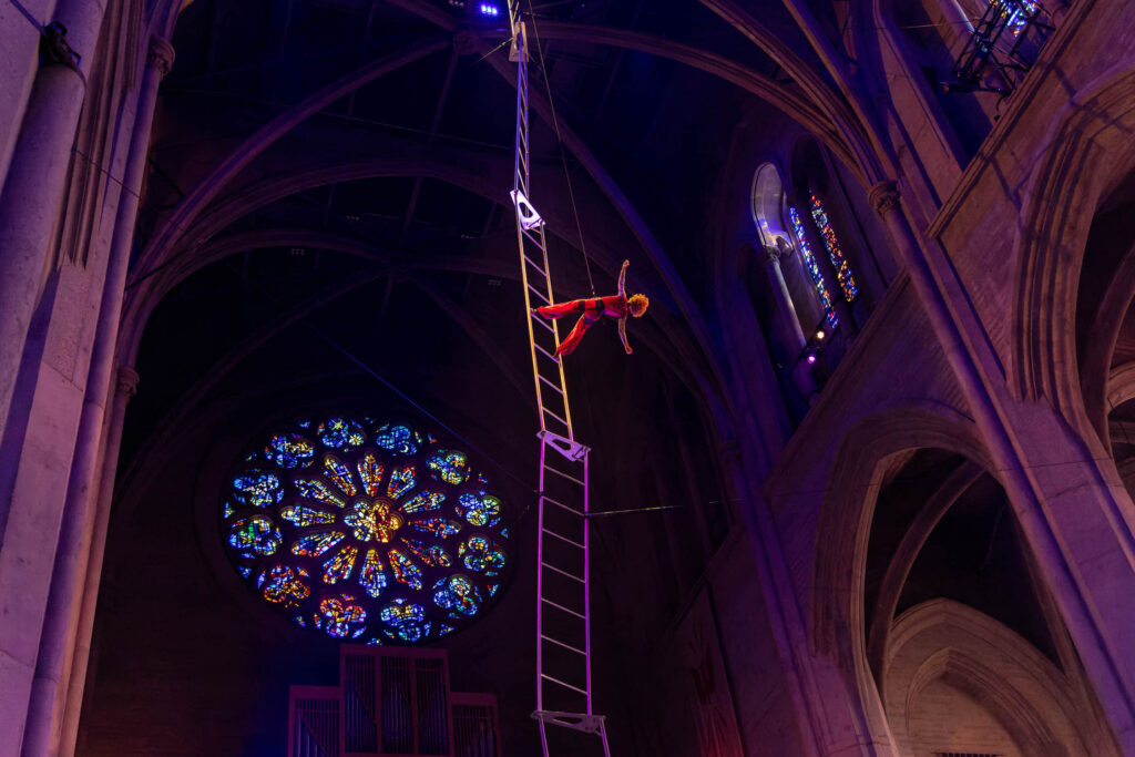 A caramel-colored Black woman hangs suspended in midair, facing upwards, attached to rigging from the ceiling of a cathedral.