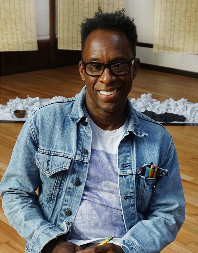 Black man up close, glasses, smiling, stack of pencils in jean jacket pocket.