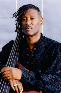 Black man with dreadlocks and mustache wearing a black and blue floral patterned blouse hands folded over an upright bass.