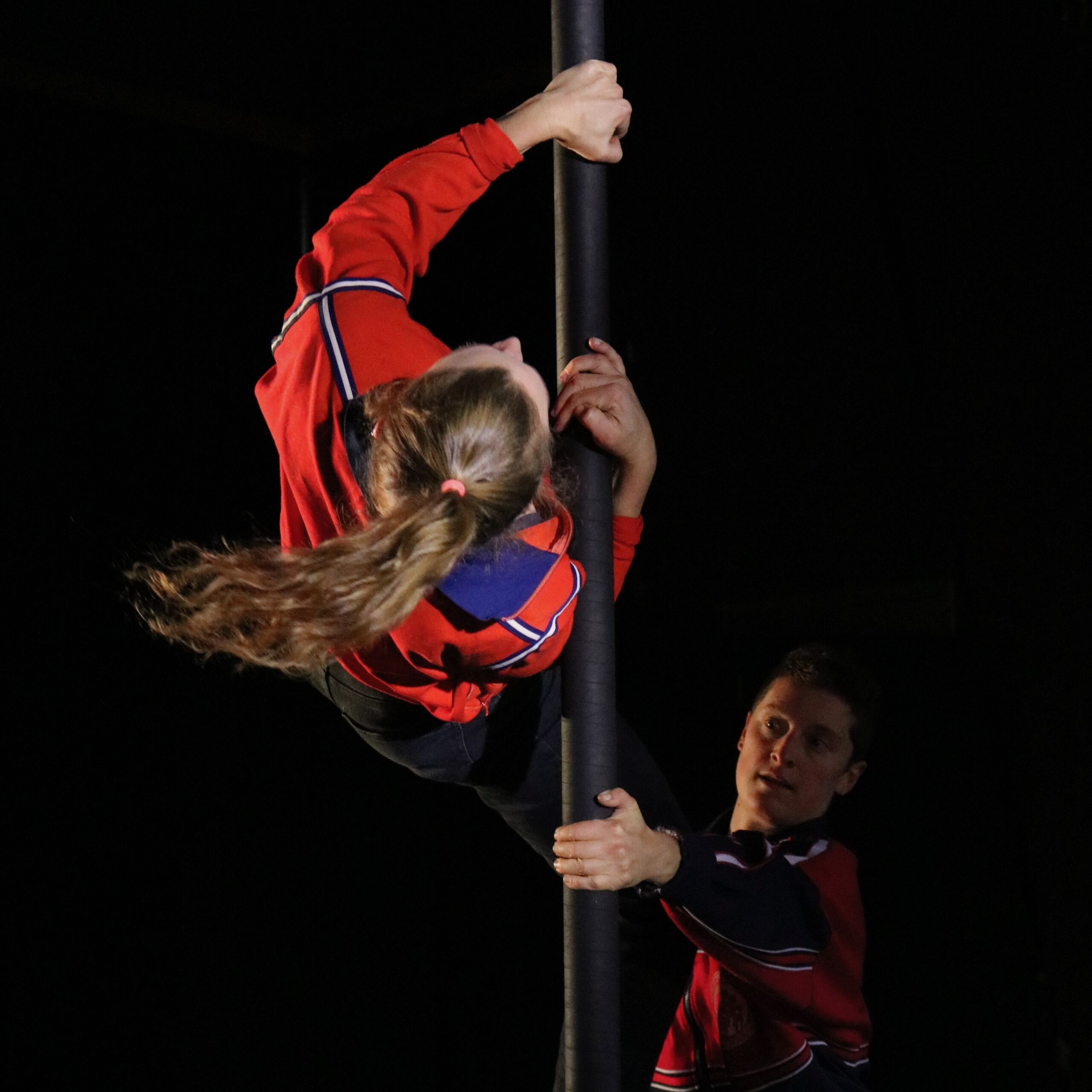An acrobat in a red jacket does a flip on a black pole, while her sister watches from the other side of the pole.