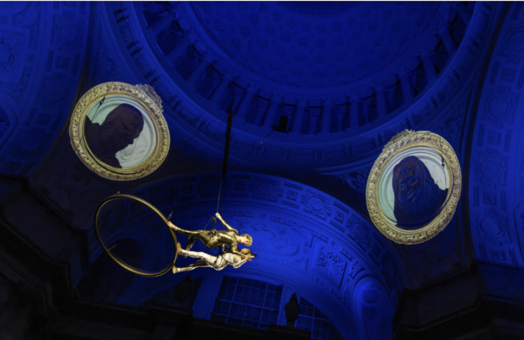 2 aerialists Veronica Blair and Ciarra D'Onofrio performing with a blue light and faces projected behind them in the rotunda of SF City Hall
