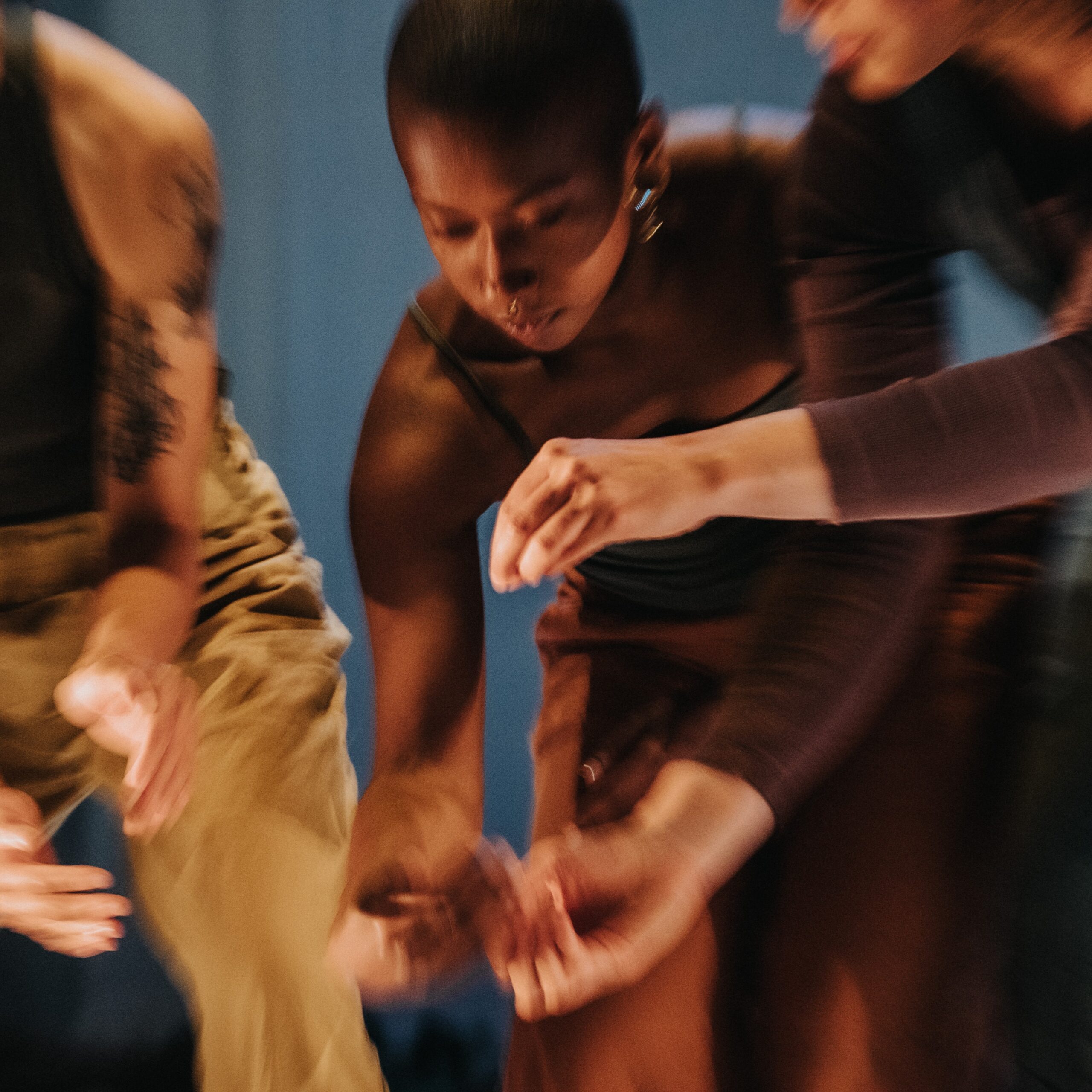 Three dancers wearing different shades of brown bend their upper bodies and bring their hands together.