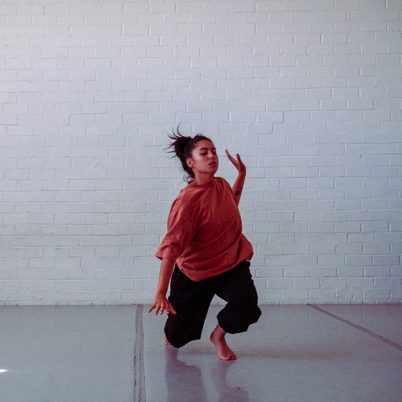 A photo of a dancer in a white studio space moving with both legs bent and left hand lifted.