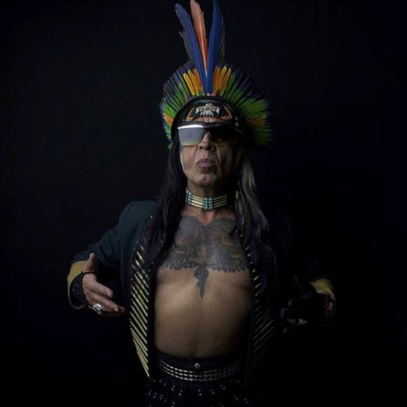 A photo of one performer wearing large feather headpiece, sunglasses and black jacket on a naked tattooed torso.