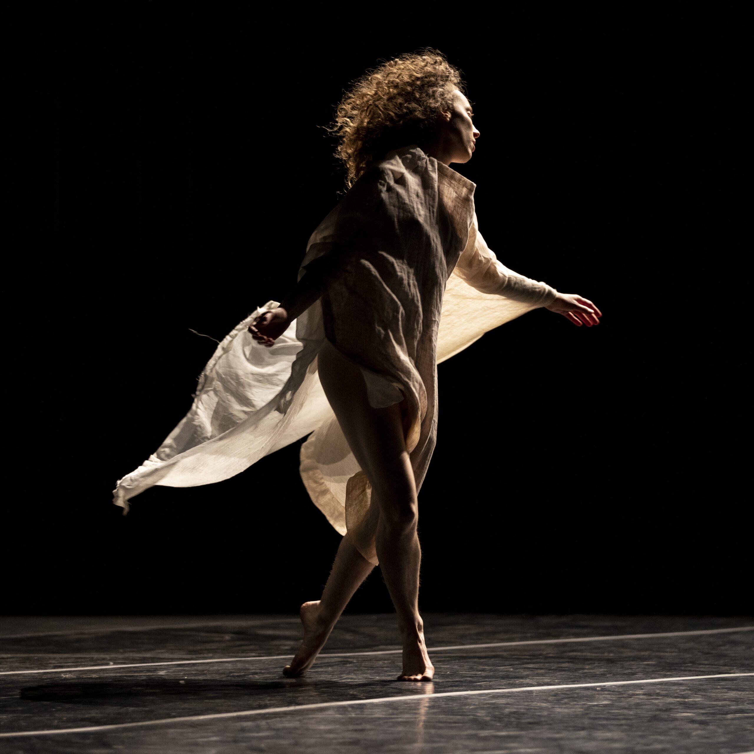 One dancer with long and curly hair wearing long flowy beige costume makes a step on stage.
