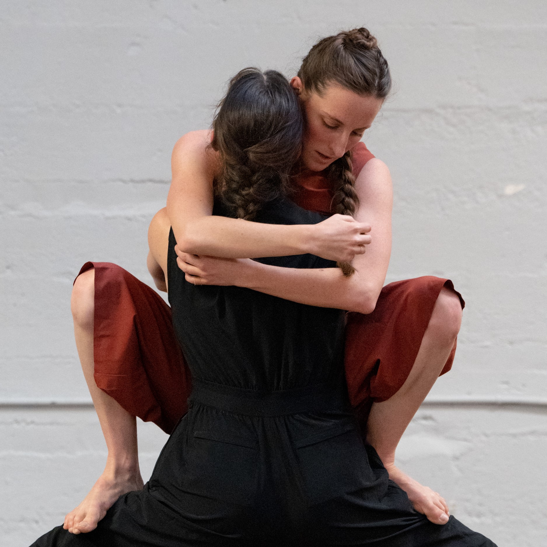Two dancers with long braided hair perform in a space with white walls. One dancer is lifting another in a hug-like movement.