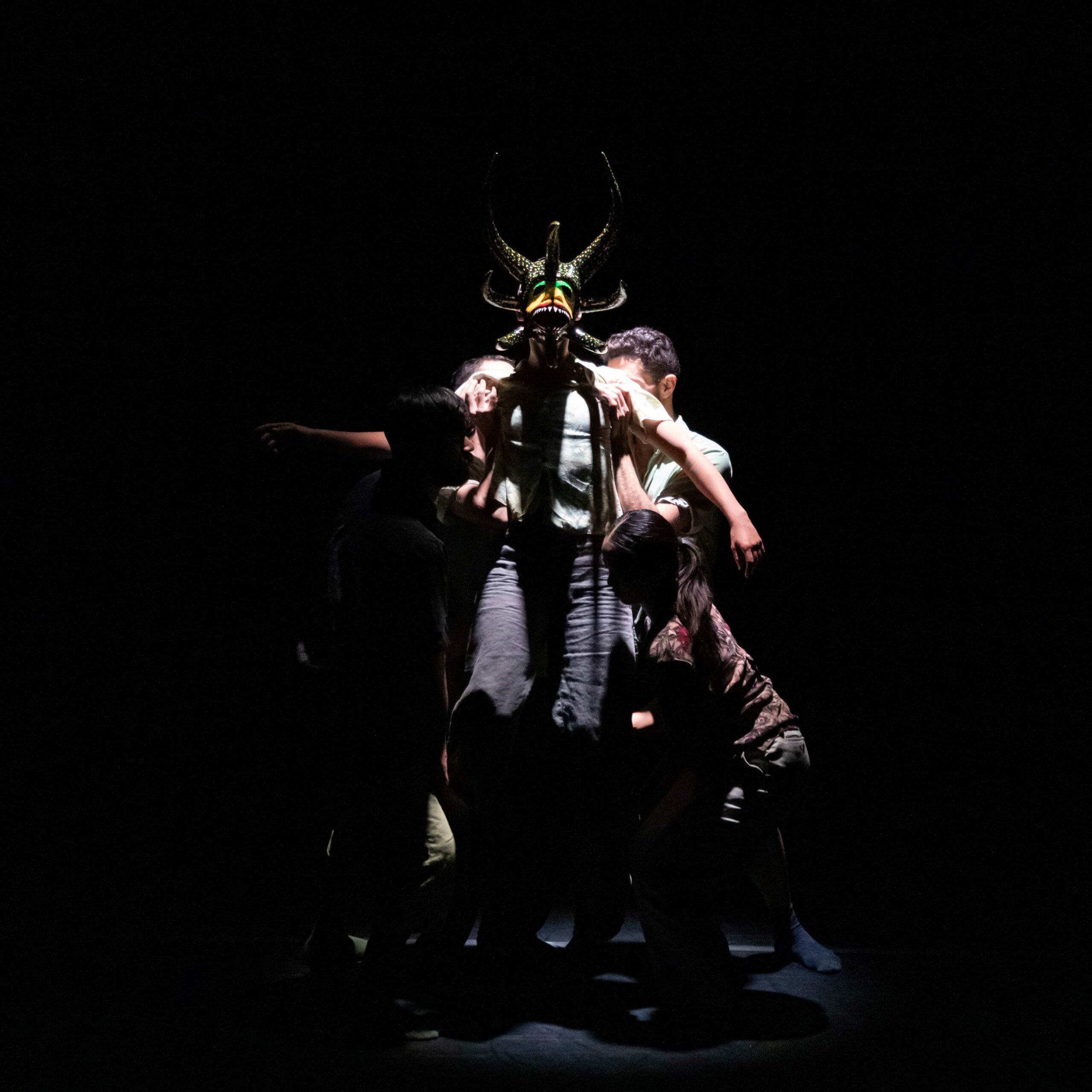 Four dancers stand in a spot light and lift another dancer wearing a big monster-like face mask.