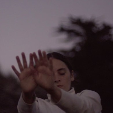 A photograph of a dancer in a white long sleeve stretching out their hands to the front.