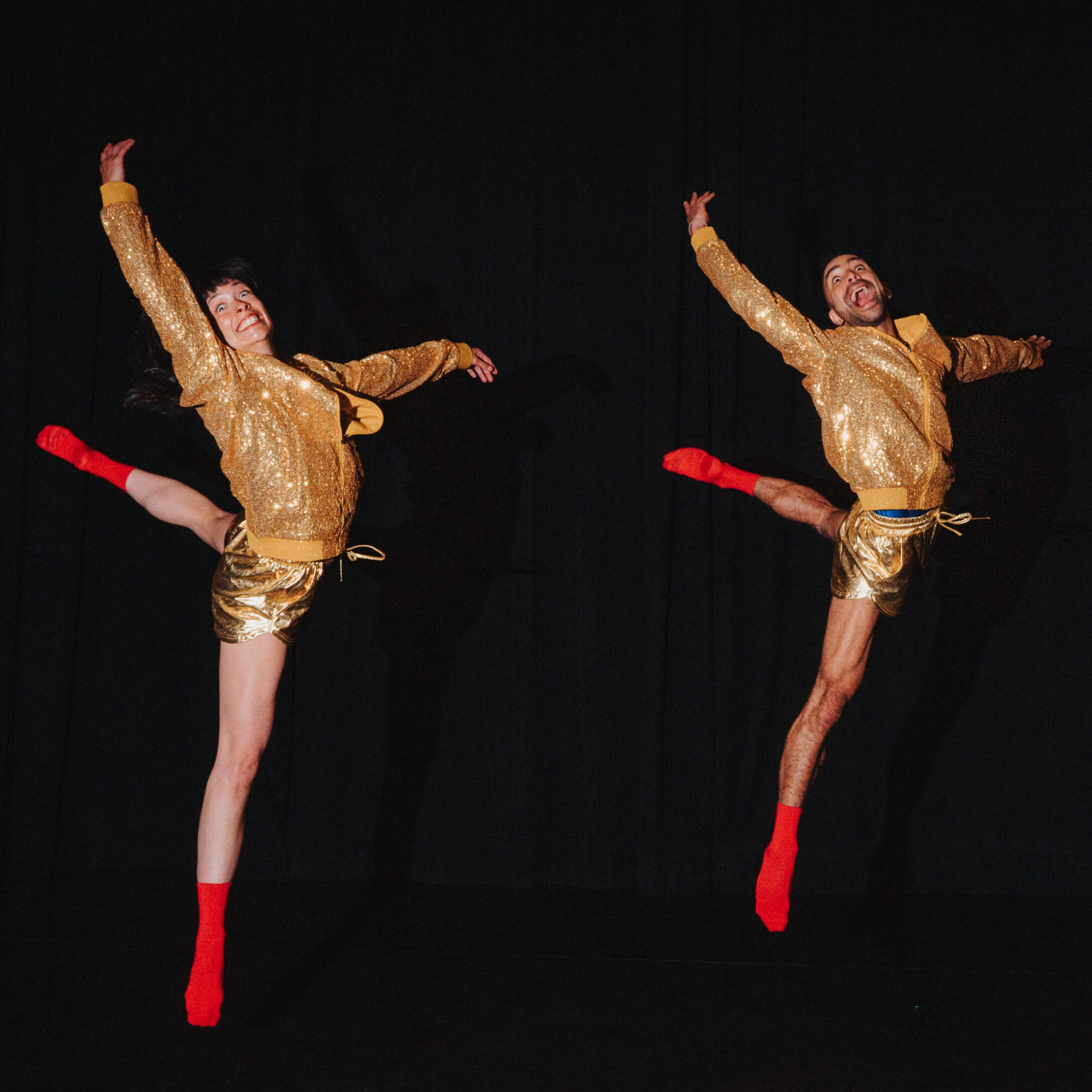 Against a black background, two dancers side by side wearing gold sparkly jackets and shorts, bare legs and red socks, jump with one leg bent one arm up and their mouths open and eyes wide