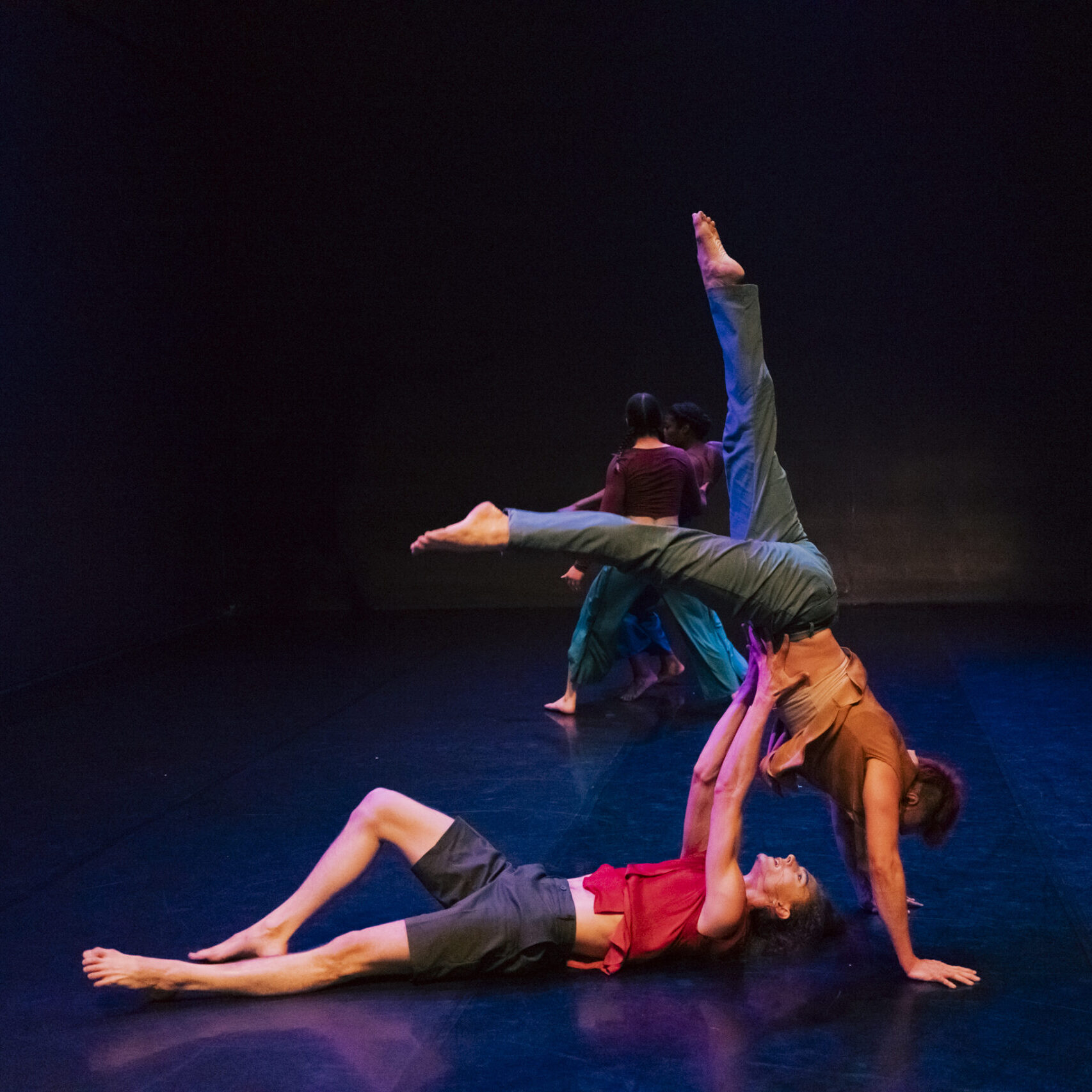 Photo of 4 dancers, in the foreground a dancer lies on their back with their arms raise supporting another dancer who is upside down in a hand stand position with their legs splayed. The lighting around the dancers is mostly black except for the light on the dancers and the floor is dark blue.