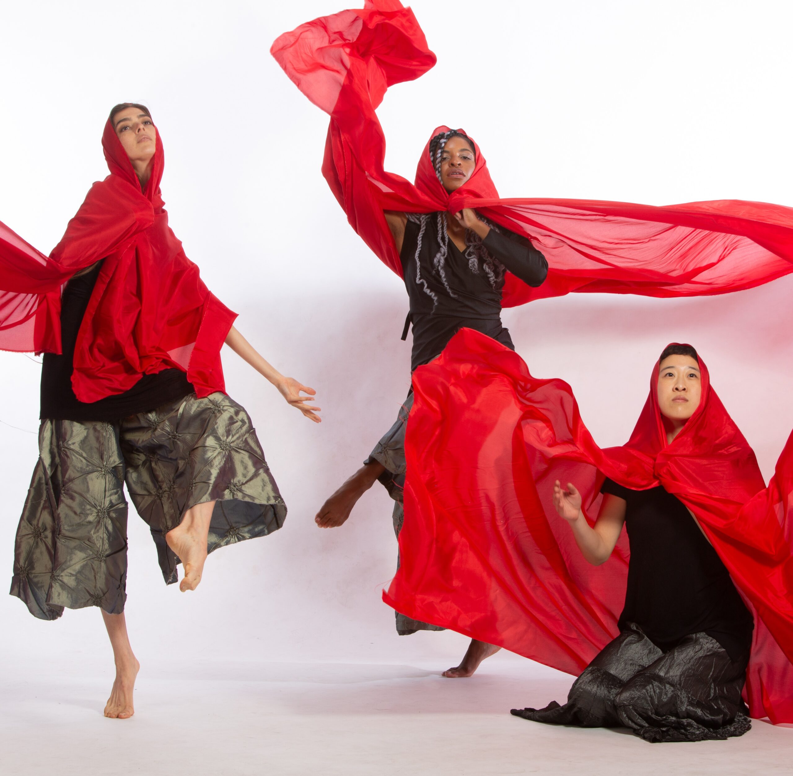 This is a photo of 3 dancers, each wearing flowy shiny dark gray/green pants and black tops. Large pieces of sheer red fabric are draped around their shoulders and heads, and floating in the air around them. The dancer on the left is up on one leg looking longingly upwards with an arm stretch downward. In the center is another dancer on one leg, staring into the camera. On the far right is a third dancer, kneeling and looking upwards with an arm outstretched.