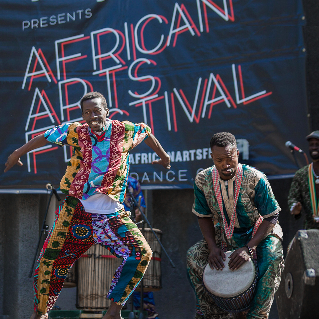 Energetic dancer in colorful outfit leaps beside a West African Drummer