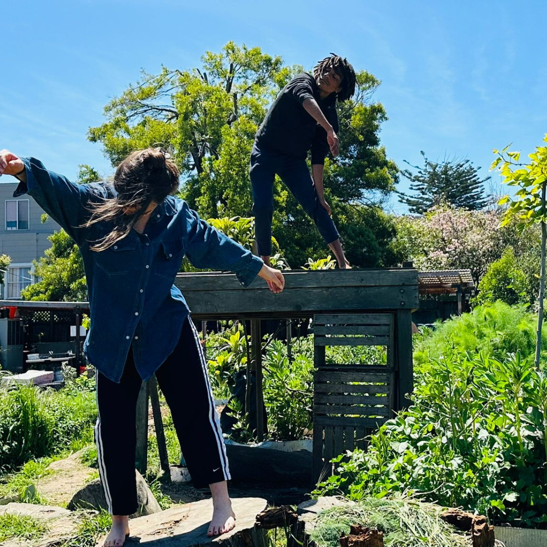 Two dancers standing, one on the ground and one on a wooden structure, and bending to one side. In their background are trees, bushes, and a bright blue sky.