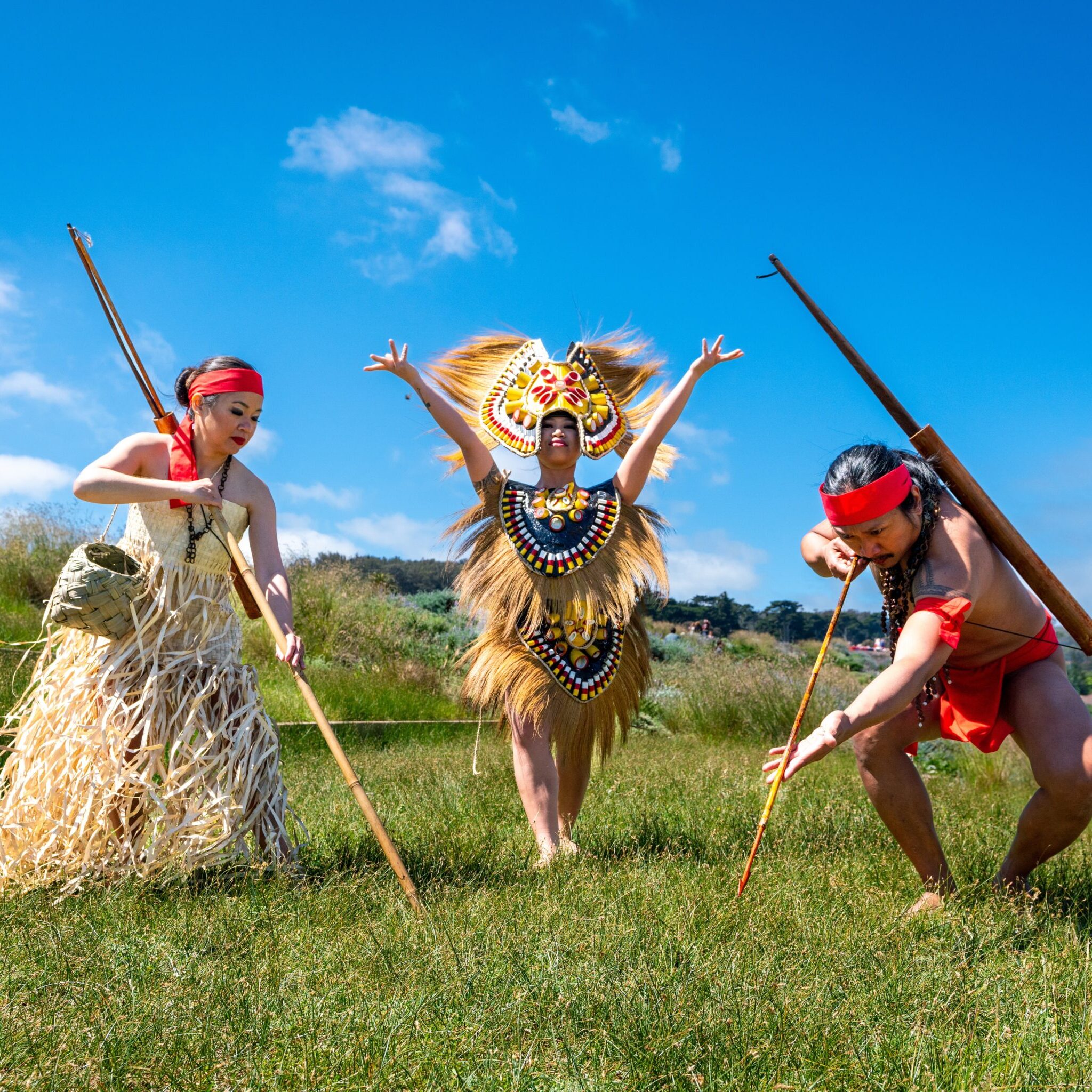 Three Filipino dancers in cultural attire and headdress. 2 women in tan grass-colored dresses, one carries a basket and large stick, the other is in a two piece ensemble with a large headdress made of grass and yellow/blue/red materials. The male dancer is wearing a red headband and loincloth while posing with a traditional bow and arrow. 