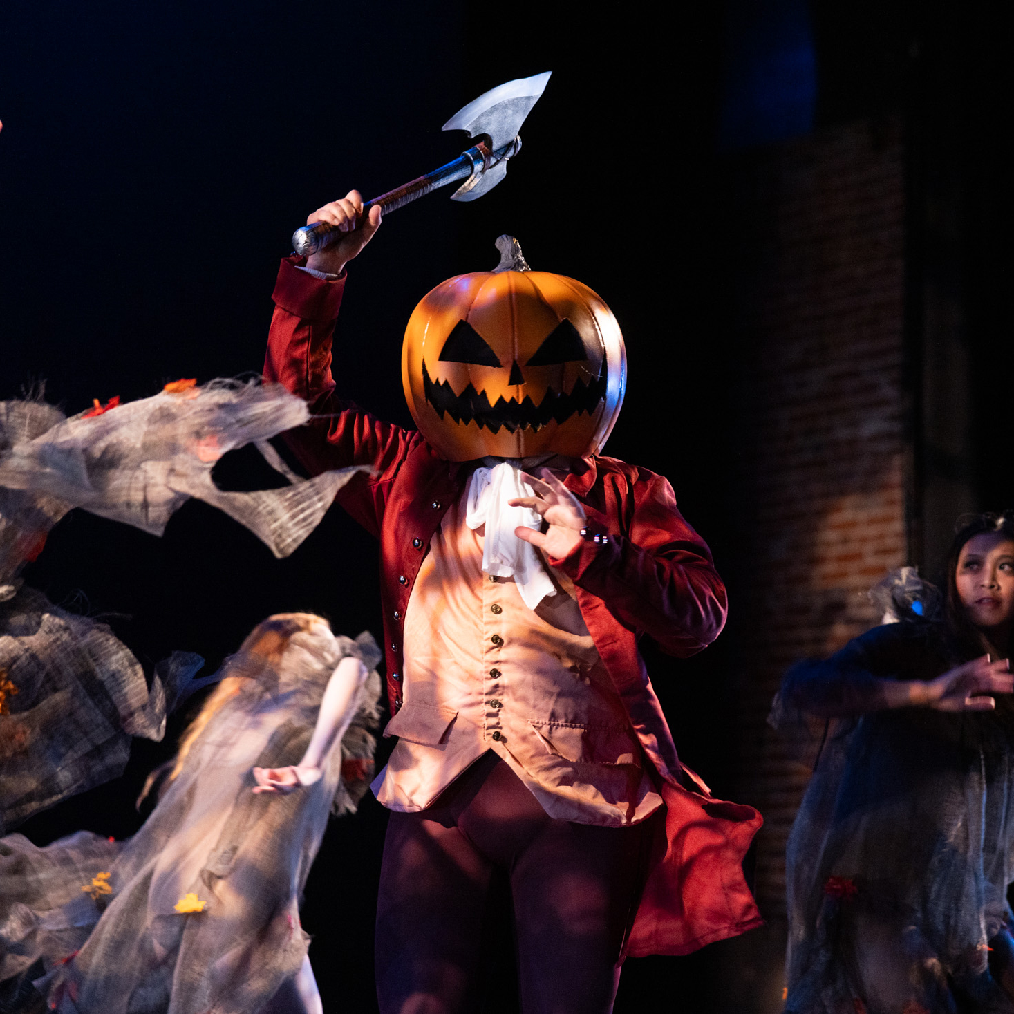 A dancer in a coat and jack-o-lantern mask raises an axe overhead as dancers in gray nightgowns swirl around him under dappled lighting.