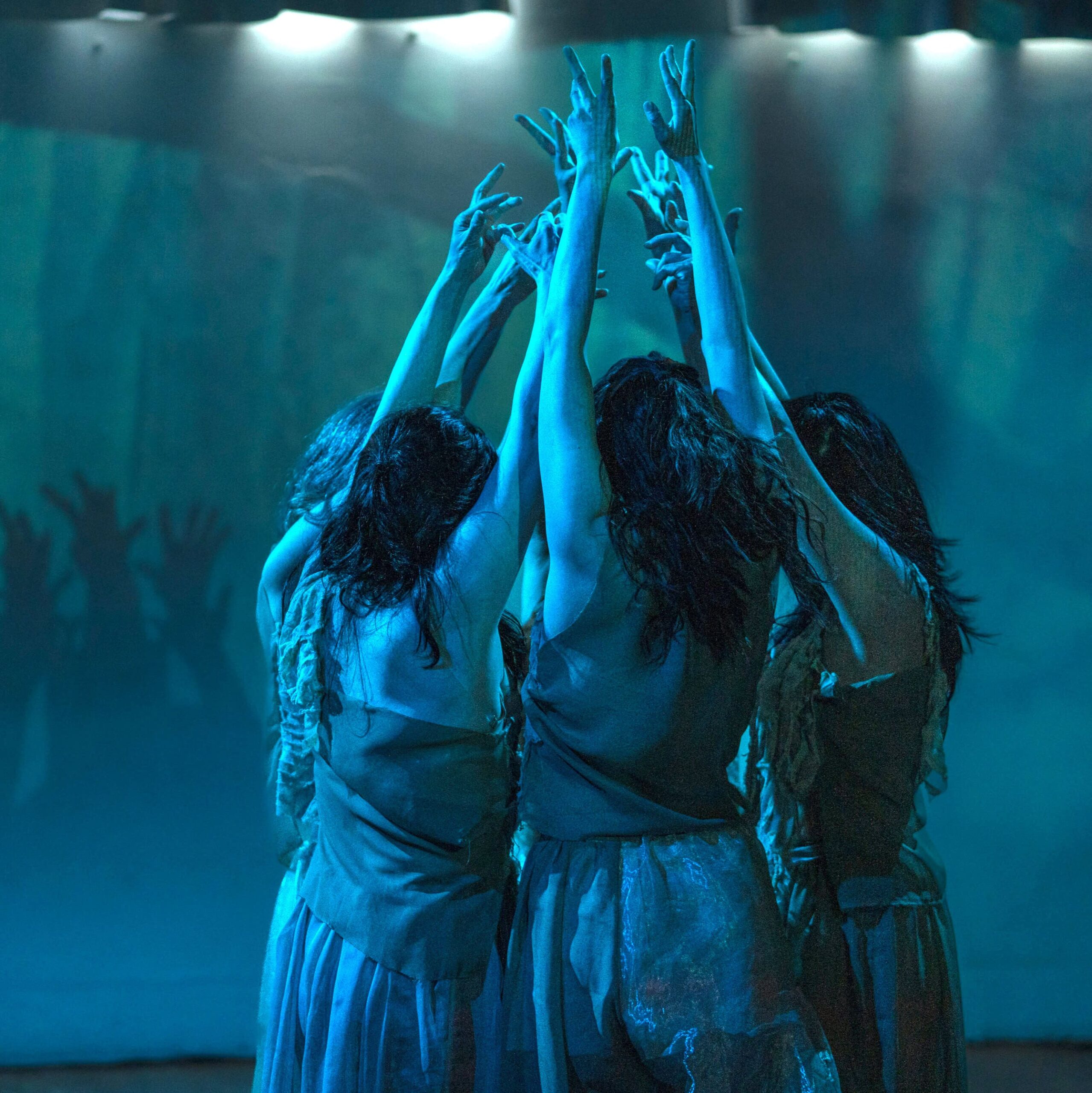 five woman wear thin fabric with no sleeve top and skirt, facing each other gathering tight reach with both arms to the celling in the blue light. 