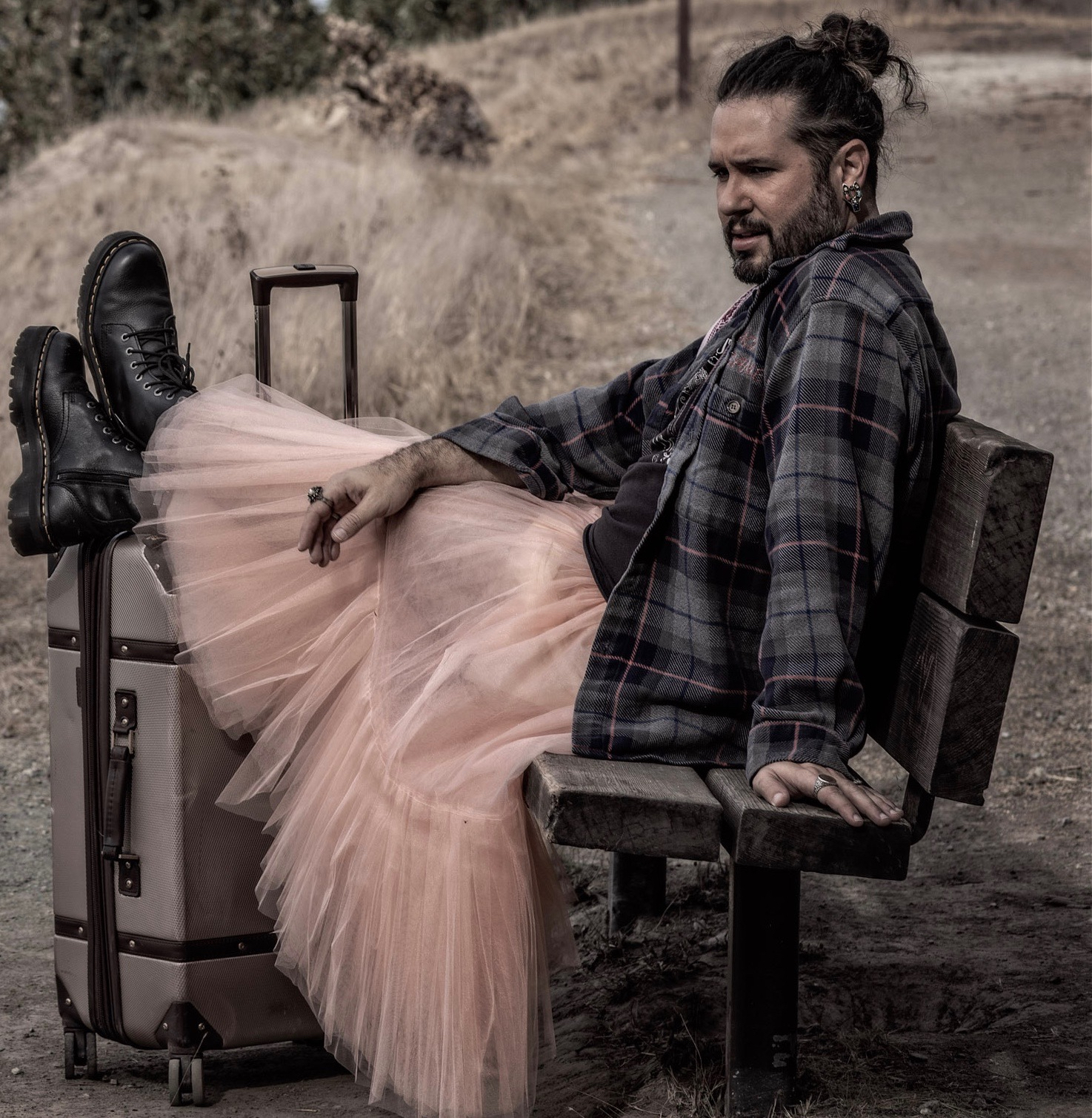 A dancer sits outdoors on a park bench, their feet propped on a suitcase. They wear a flannel long sleeved shirt and a pale pink long tulle skirt.