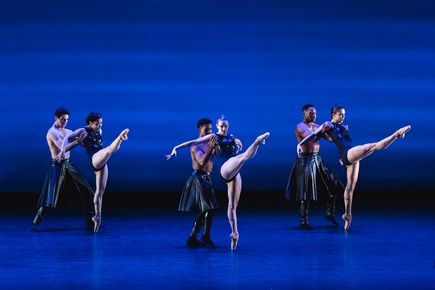 A performance photo of six dancers, in three pairs, against a blue-lit background.