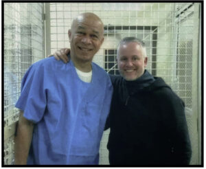 Two men stand in a small white cage used for prison visitations with their arms around each other, smiling and facing the camera.