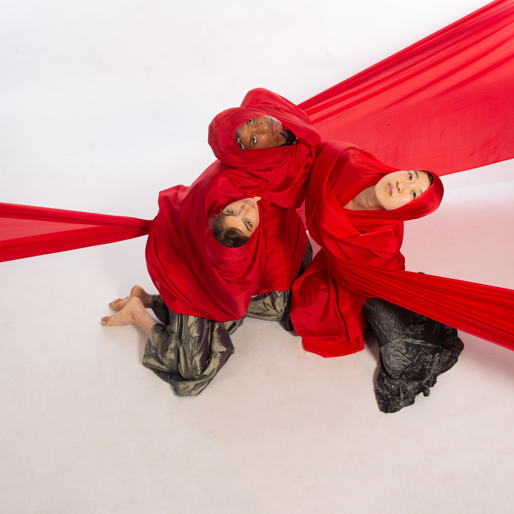 Three women seated in a circle look up at the camera. Dressed in black tops with metallic pants, they wrap red silk around their shoulders and head. 