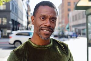 A handsome Black man with a short, neat haircut, smiling with head slightly tilted, wearing a green velvet shirt.