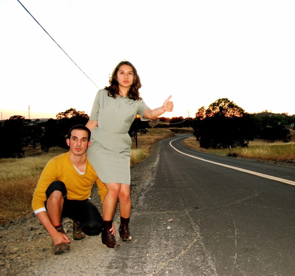 Eric Garcia and Kat Cole hitchhiking on the side of a country road at sunset.