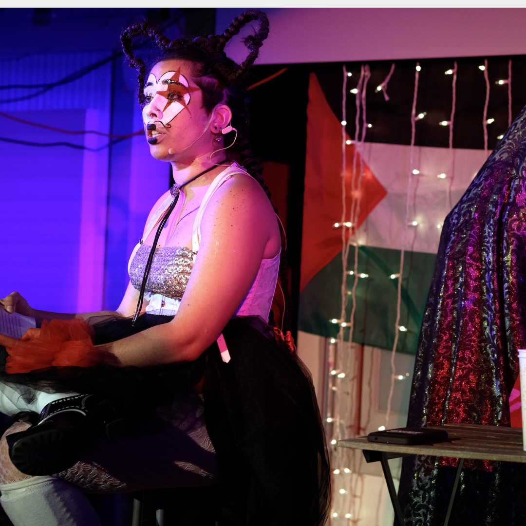 Image of Mylo Cardona (they/them) performing with a painted face, hair in braids above their head, sitting on a stool. Behind them is a Palestinian flag