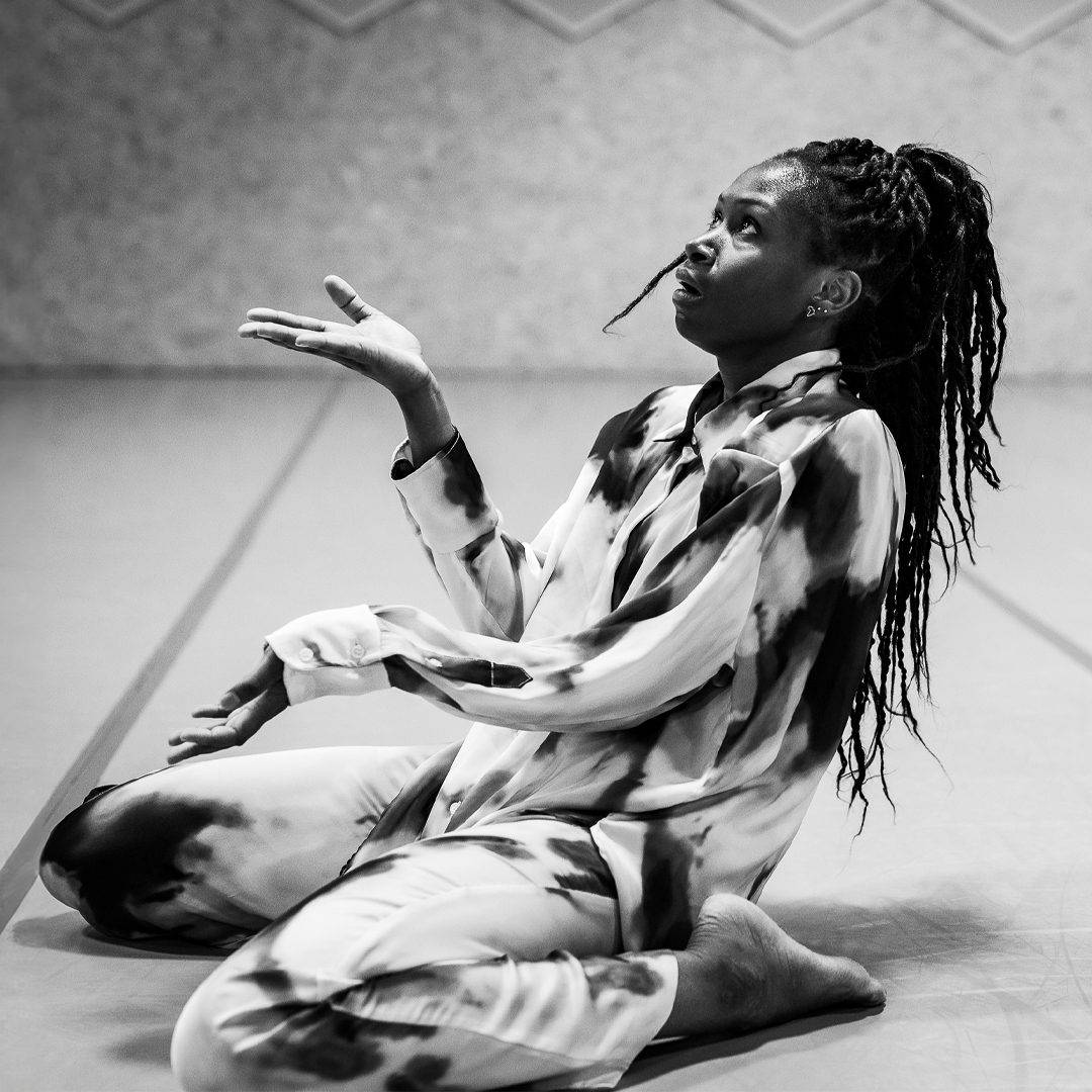 A  black and white photo of a kneeling woman sitting on her heels. She is slightly leaning backwards with her torso. Her head is tilted towards the sky. Her left arm is extended in front of her with an open upward facing palm. Her right arm in front of her is bent at the elbow with an open upward facing palm. She seems to be expecting and hoping for something to fall from the sky into her hands. 