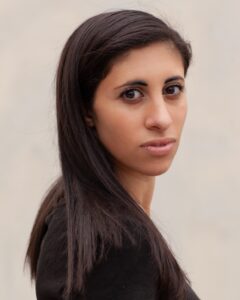 Leila, an Arab woman (she/her) stands against a beige backdrop and looks toward the viewer. 