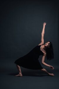 A dancer with long black hair in a dark blue dress is in a lunge with the right leg in front and right arm up, leaning backwards.