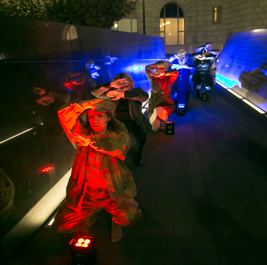 Six dancers are kneeling in a staggered row with their arms folded near their face. They are in between two concrete walls and blue and red lights are being cast onto the dancers.