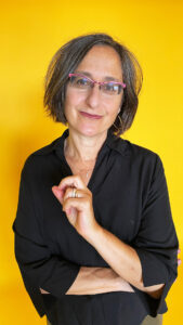 A middle aged white cis woman with a brown/gray bob in a black shirt and pink eyeglasses smiles toward the camera.