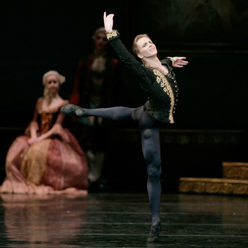 Dancer Tiit Helimets performs as Siegfried in Swan Lake. He is on a stage in a back attitude position and wears a black costume with gold accents.