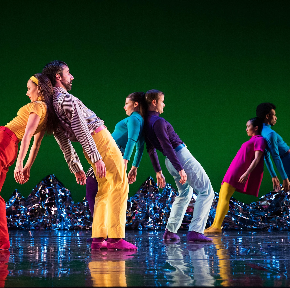 Dancers dressed in colorful retro clothing lean against each other in front of a green background.