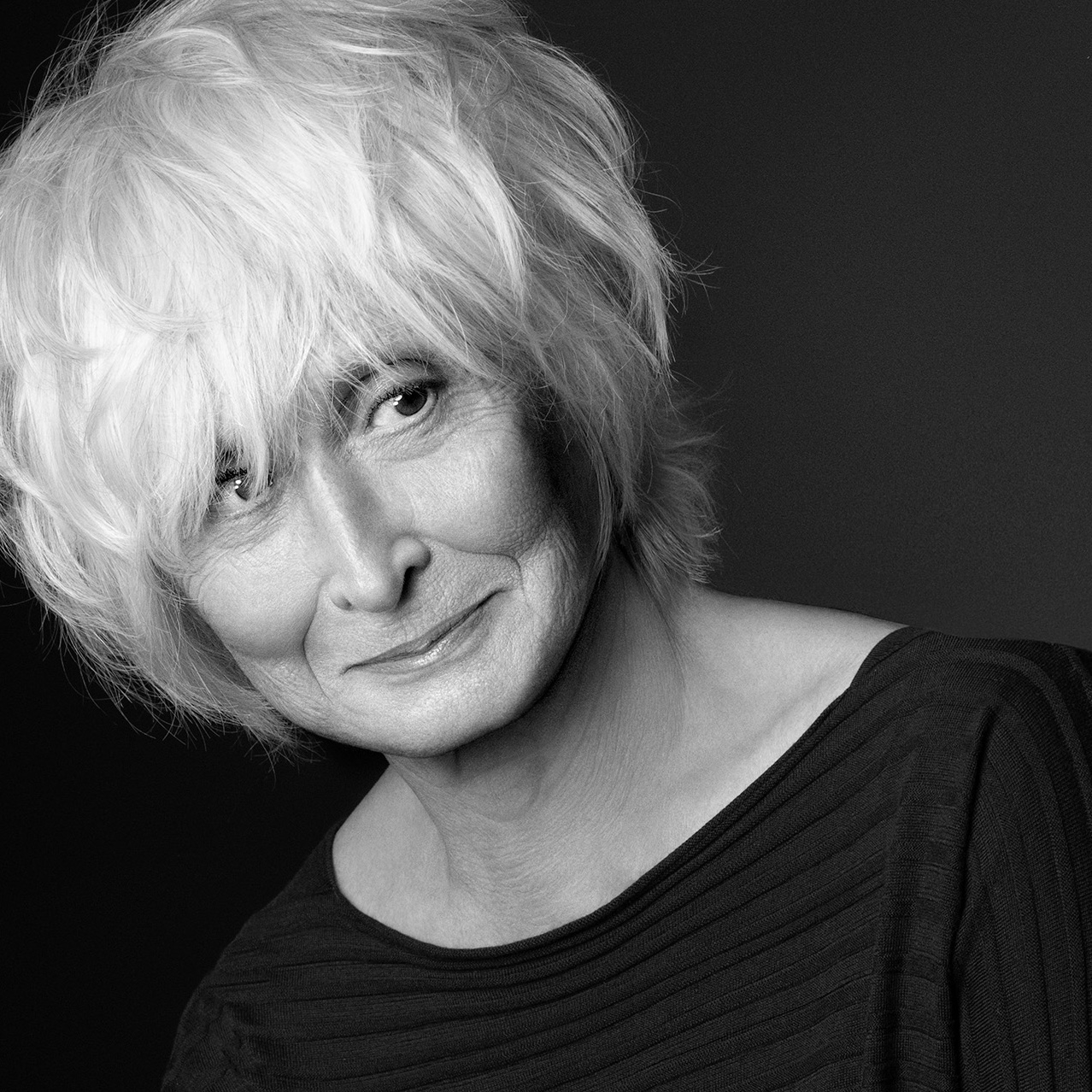 A black and white image of Twyla Tharp in front of a dark background.