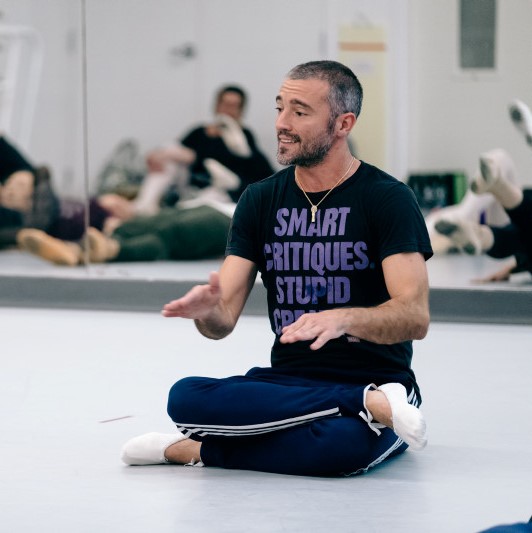 A photo of a man sitting cross-legged on the floor of a ballet studio.