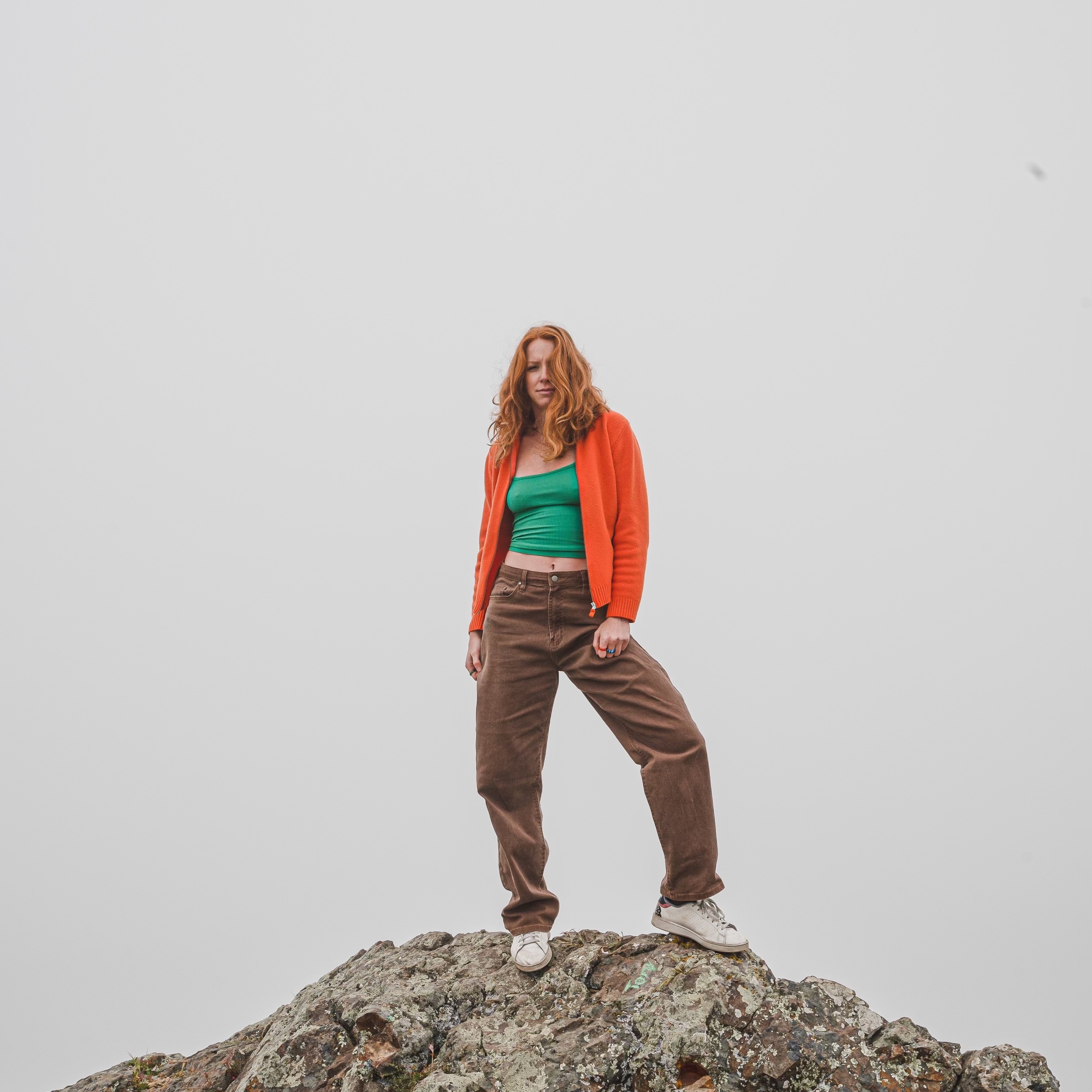 Abigail stands on rock looking outward with an open stance wearing a bright orange sweater, green shirt, and baggy brown pants.