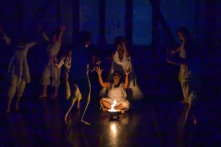 Dancers are in white. One woman presenting dancer sits in front of a flame while other dancers run around her