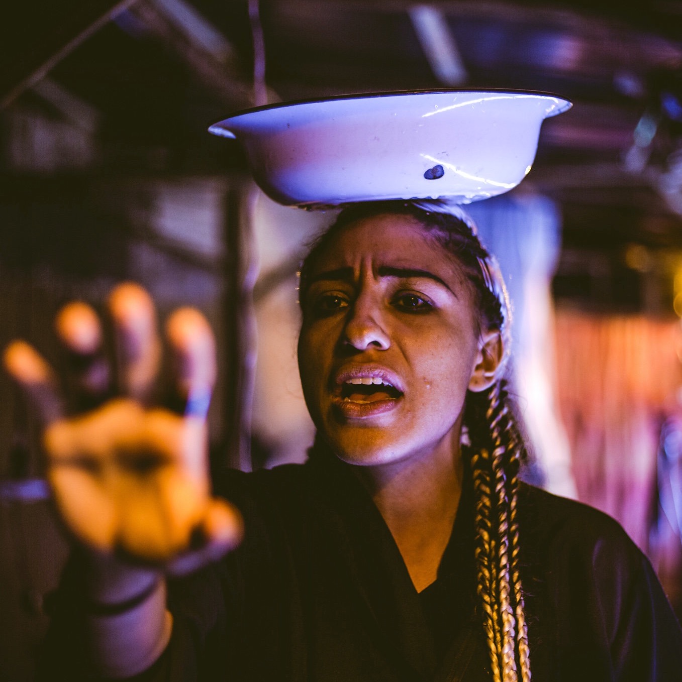 A dance artist is pictured from the torso up reaching out their right arm. They have light brown skin and they are balancing  a white bowl on her head with water in it. There are long braids cascading down their left shoulder and waist and they appear to be calling out.