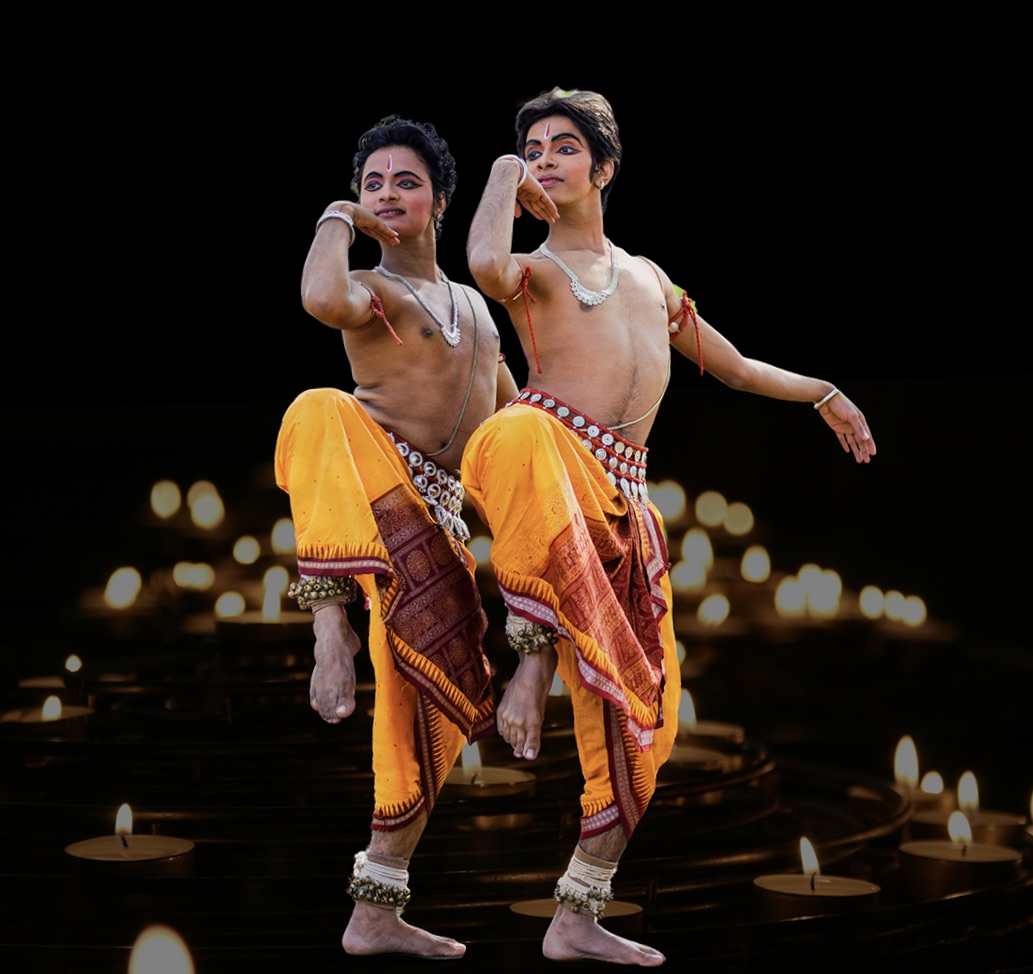 A photo two male dancers in Odissi dance costumes against a black backdrop with shimmering lights. 